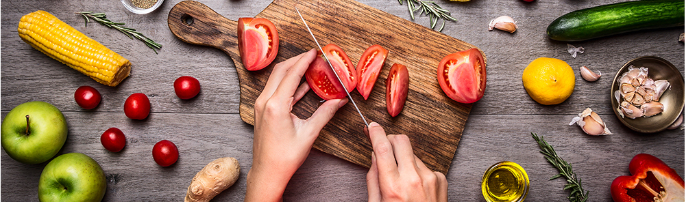 Slicing fresh tomatoes on a wooden cutting board surrounded by other fresh vegetables.